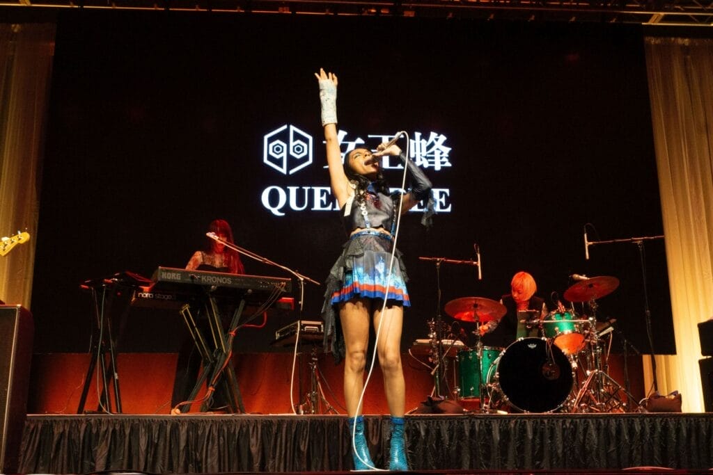 Photograph of Queen Bee performing at Anime Boston 2024. Avu-chan is dressed in a black, blue, and red outfit with sky blue boots, reaching up as she leans back and belts out a note.