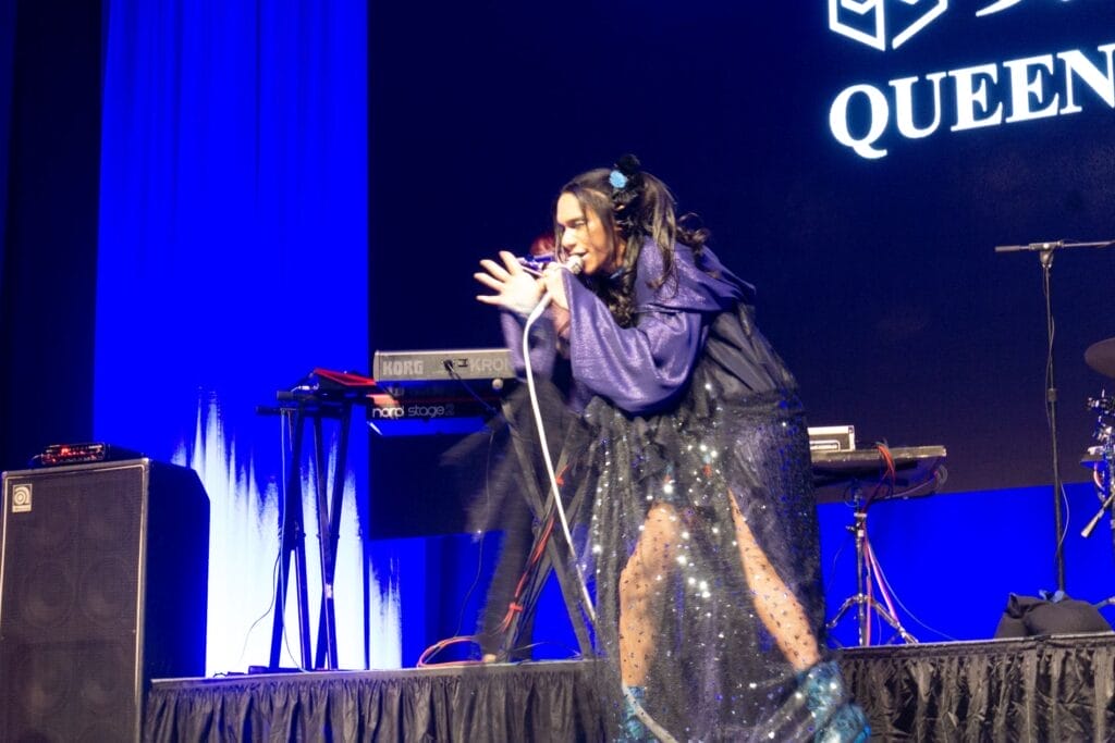Photograph of Queen Bee performing at Anime Boston 2024. Avu-chan is dressed in a black and purple cloak as she sings.