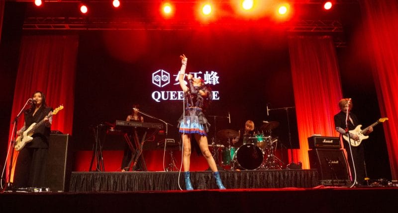 Photograph of Queen Bee performing at Anime Boston 2024. Avu-chan is dressed in a black, blue, and red outfit with sky blue boots, standing straight as they sing. Hibari-kun, meanwhile, plays the guitar while wearing a black, short-sleeved shirt and Yashi-chan plays bass wearing a black sleeveless shirt