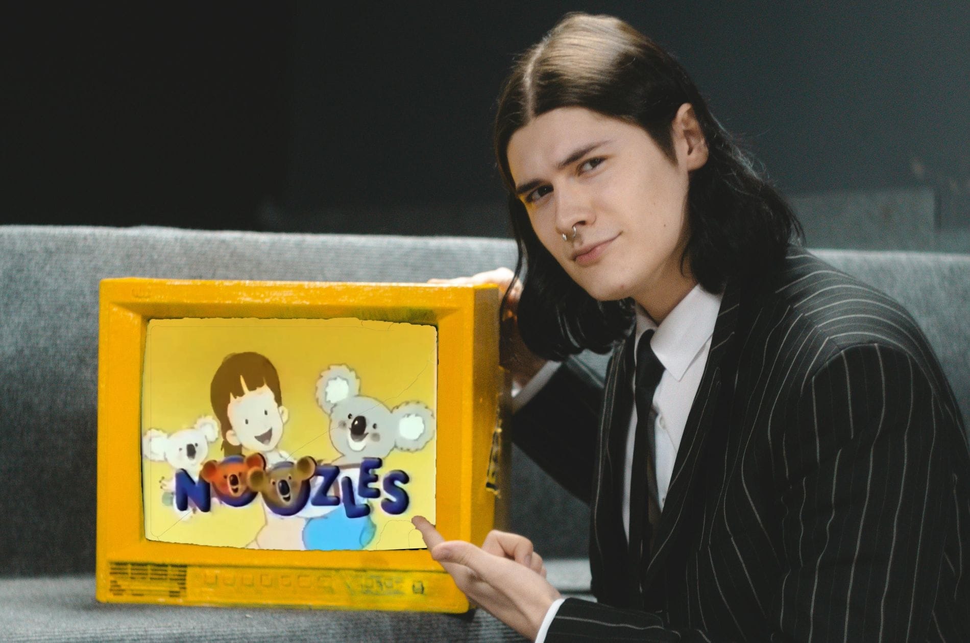 A man in a suit looks seriously as he points toward a yellow TV that's playing Noozles.