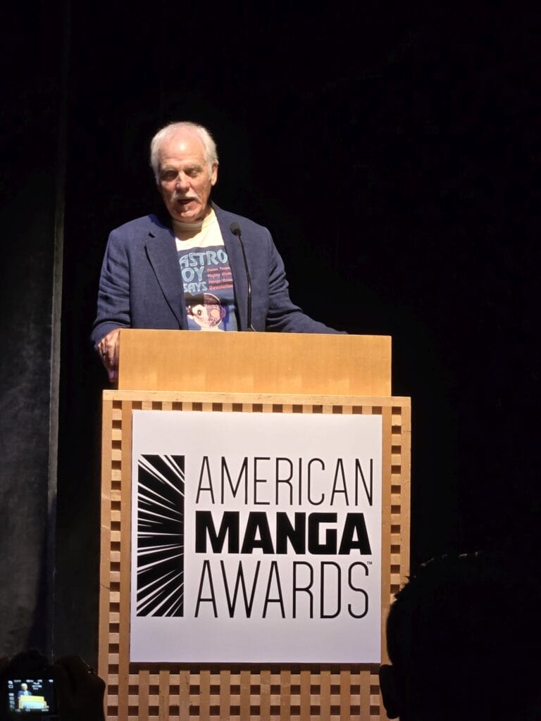 Frederik Schodt speaking at a lectern at the American Anime Awards. He is wearing a blazer and an Astro Boy T-shirt.