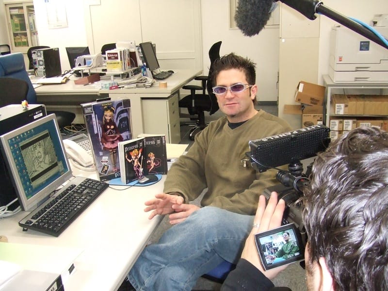 A photograph of Stu Levy, a man wearing Oakley sunglasses, an olive sweater, and blue jeans. He's sitting at at a desk, which has a laptop and several Princess Ai merchandise items set up on it