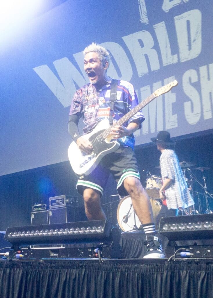 Photograph of J-Rock group FLOW's guitarist Takeshi Asakawa performing onstage against a backdrop that reads "FLOW World Tour: Anime Shibari.