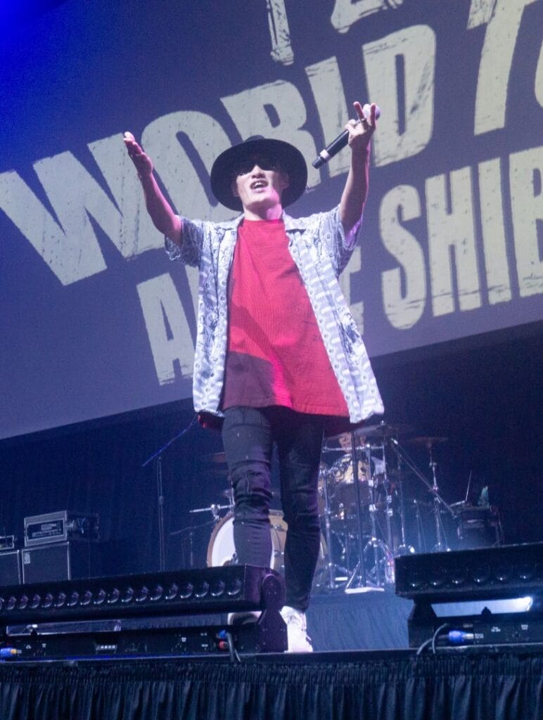 Photograph of J-Rock group FLOW's vocalist Kōshi Asakawa performing onstage against a backdrop that reads "FLOW World Tour: Anime Shibari.