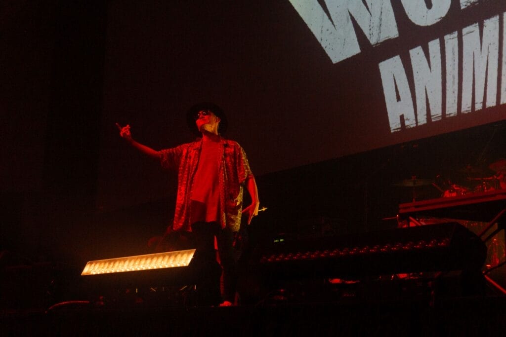 Photograph of J-Rock group FLOW's vocalist Kōshi Asakawa performing onstage against a backdrop that reads "FLOW World Tour: Anime Shibari. He's bathed in a harsh red light as he smirks