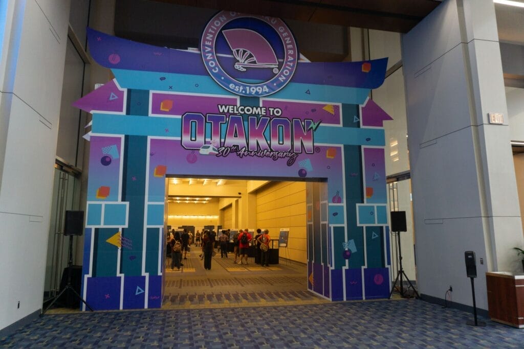 Photograph of a large, cardboard, blue and purple tori gate with a sin that reads "Welcome to Otakon 30th Anniversary"