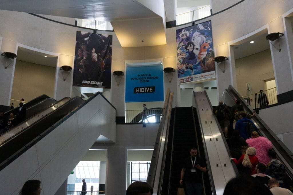 Photograph of people riding up an escalator. At the top, three posters are hung:
Left: Wind Breaker anime
Middle: A blue banner that reads "Have a Wicked Good Con! - HIDIVE"
Right: Advertisement for the Mono anime