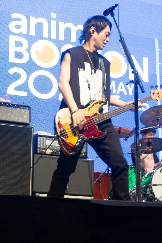 Photograph of Yuuta Tsujimura from Blue Encount. He's wearing a sleeveless black t-shirt as he plays the bass in front of a blue sign that reads "Anime Boston 2025"