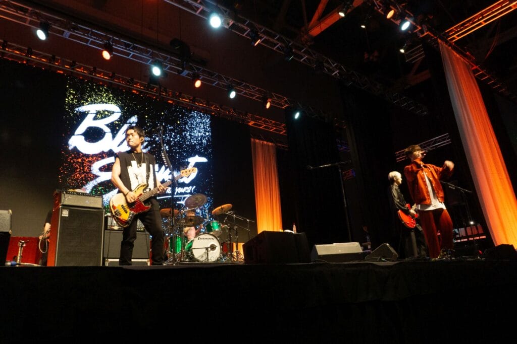 Photograph of Blue Encount playing onstage in front of a lighted sign with the band's name on it.