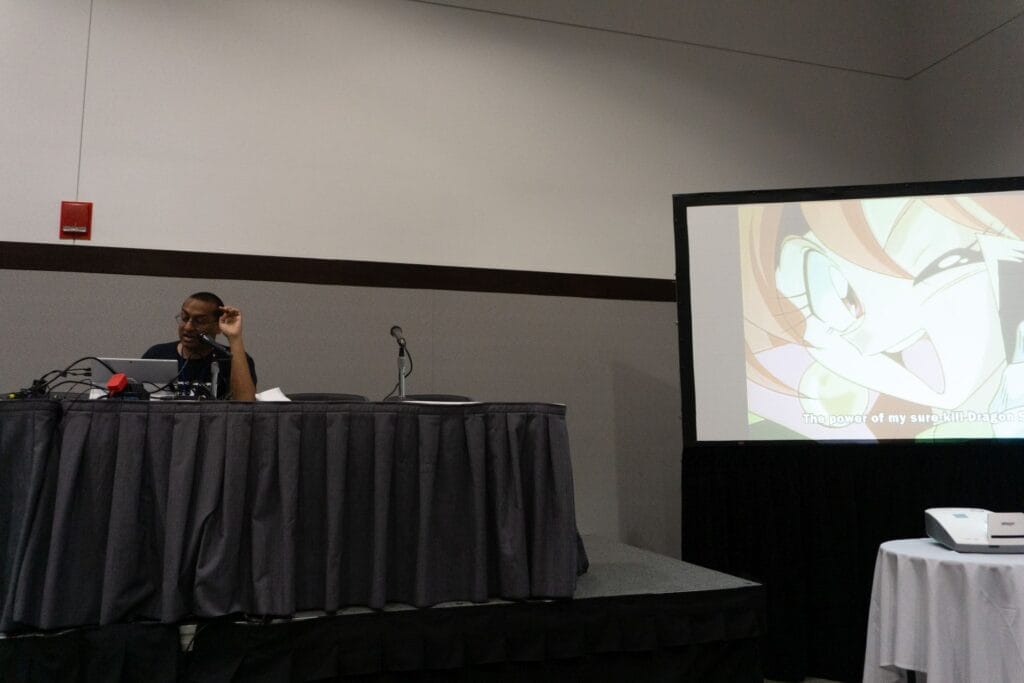Photograph of Daryl Surat at Anime Boston 2025, sitting next to a screen depicting Lina Inverse