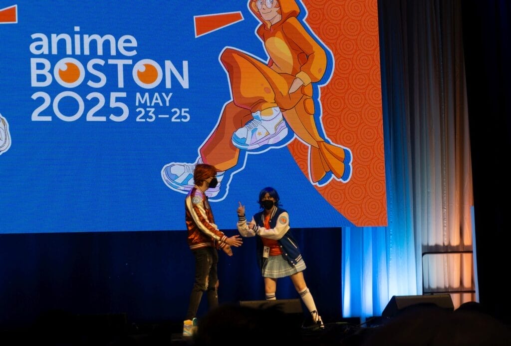 Photograph from Anime Boston 2025's opening ceremonies of A-chan and B-kun, an orange-haired man and a blue-haired woman, respectively, wearing casual clothes and talking with each other onstage.