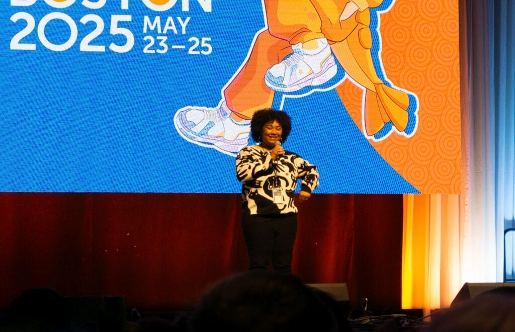 Photograph from Anime Boston 2025's opening ceremonies of Anairis Quinones smiling, hand on hip, as she looks at the audience.