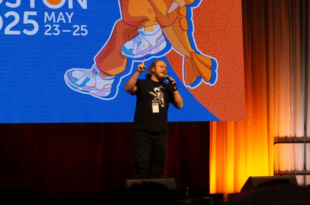 Photograph from Anime Boston 2025's opening ceremonies of Bryson Baugus excitedly talking into a microphone as he points up