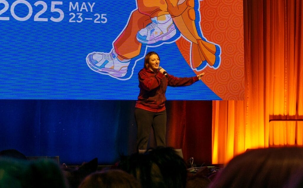Photograph from Anime Boston 2025's opening ceremonies of Caitlin Glass talking into the audience, her free arm outstretched to her side.