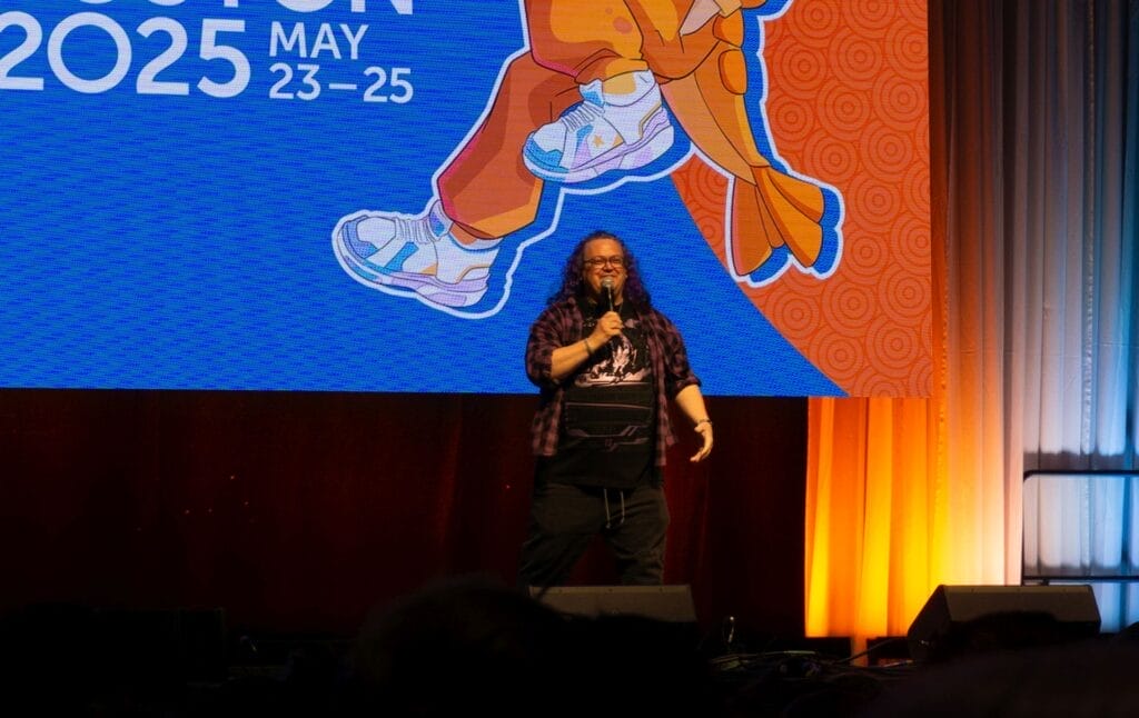 Photograph from Anime Boston 2025's opening ceremonies of Greg Ayres casually addressing the audience onstage.