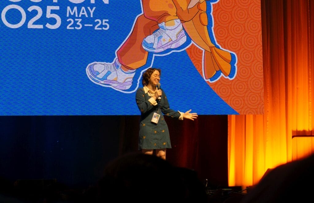 Photograph from Anime Boston 2025's opening ceremonies of Jill Harris excitedly addressing the audience. She's wearing a blue dress and glasses.