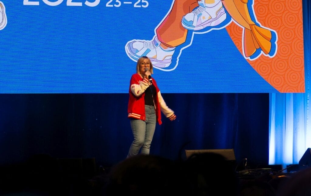 Photograph from Anime Boston 2025's opening ceremonies of Juliet Simmons talking into a microphone. She's wearing jeans and a red-and-white varsity jacket.