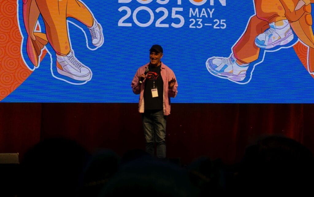 Photograph from Anime Boston 2025's opening ceremonies of Kirk Thornton addressing the audience. He's wearing a pink overshirt and a black t-shirt, and wearing a baseball cap.