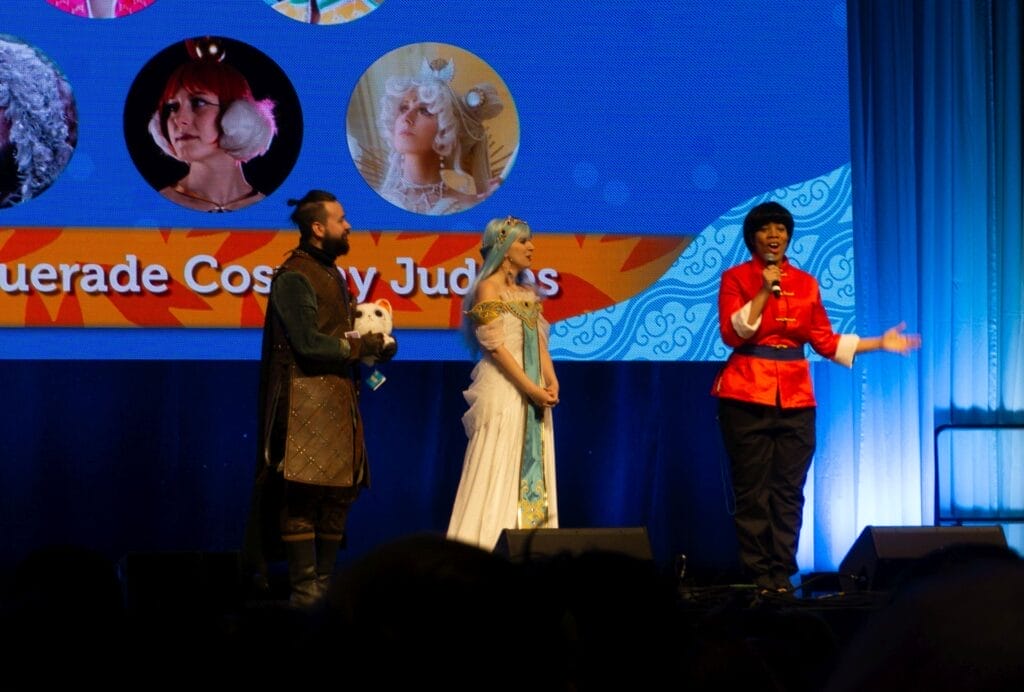 Photograph from Anime Boston 2025's opening ceremonies of three Masquerade judges. One is a Black femme individual as Ranma Saotome (Male), one is a white femme person dressed in a white gown and blue wig, and one is a white masculine individual wearing a tabard and holding a plushie.