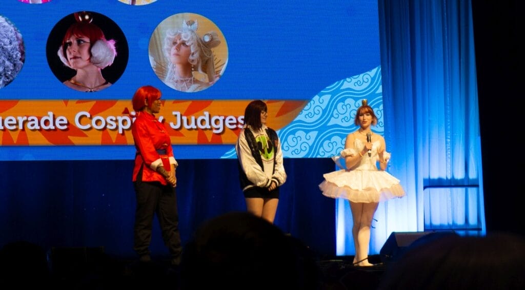 Photograph from Anime Boston 2025's opening ceremonies of three Masquerade judges. One is a Black femme individual as Ranma Saotome (Female), one is a white femme person dressed as Princess Tutu's Ahiru, And one is a femme individual dressed casually