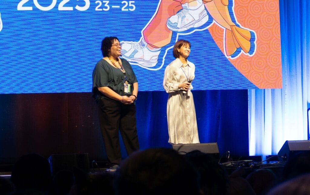 Photograph from Anime Boston 2025's opening ceremonies of Yuriko Yamaguchi, smiling as she holds a microphone. The translator is smiling and looking at her.