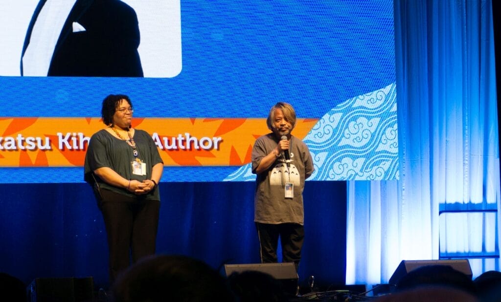 Photograph from Anime Boston 2025's opening ceremonies of Hirokatsu Kihara calmly addressing the audience. He's holding a microphone and wearing a Totoro T-shirt.