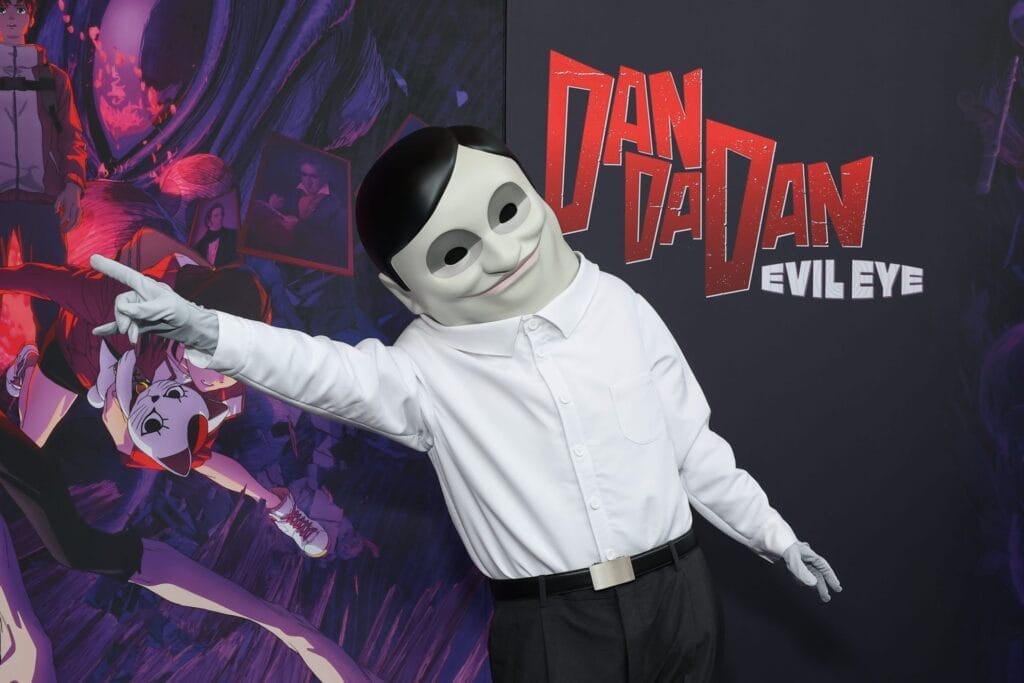 Dan Da Dan: Evil Eye world premiere that depicts a man dressed an alien - they're wearing a white mask that looks like an unnaturally man with hollow eyes and a disturbing grin, a white dress shirt, and black slacks. They're posing in front of a sign with the show's logo.
