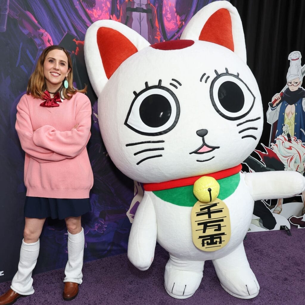 Photograph taken at the Dan Da Dan World Premiere which depicts Abby Trott, who is wearing a loose pink sweater, smiling as she poses next to a giant mascot dressed like a lucky cat.
