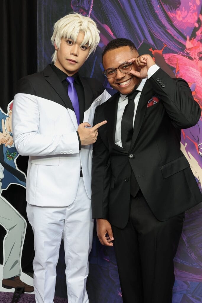 Photograph of Aleks Lee and AJ Beckles smiling as they pose next to each other. Aleks Lee is wearing a black-and-white suit with a purple tie, while AJ Beckles is wearing a black suit and tie with a red pocket square.