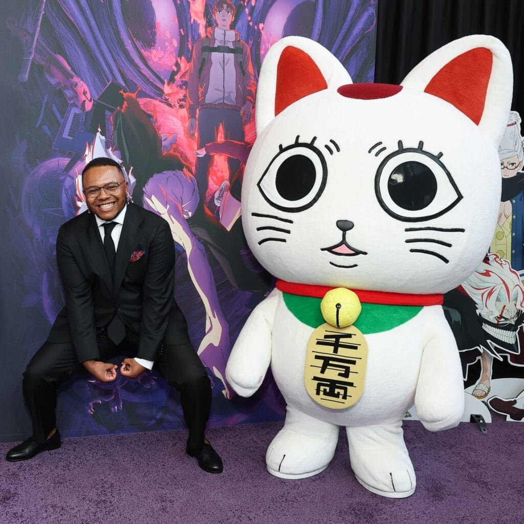 Photograph of AJ Beckles smiling as he poses next to a mascot dressed as a lucky cat. He's wearing a black suit and tie with a red pocket square.