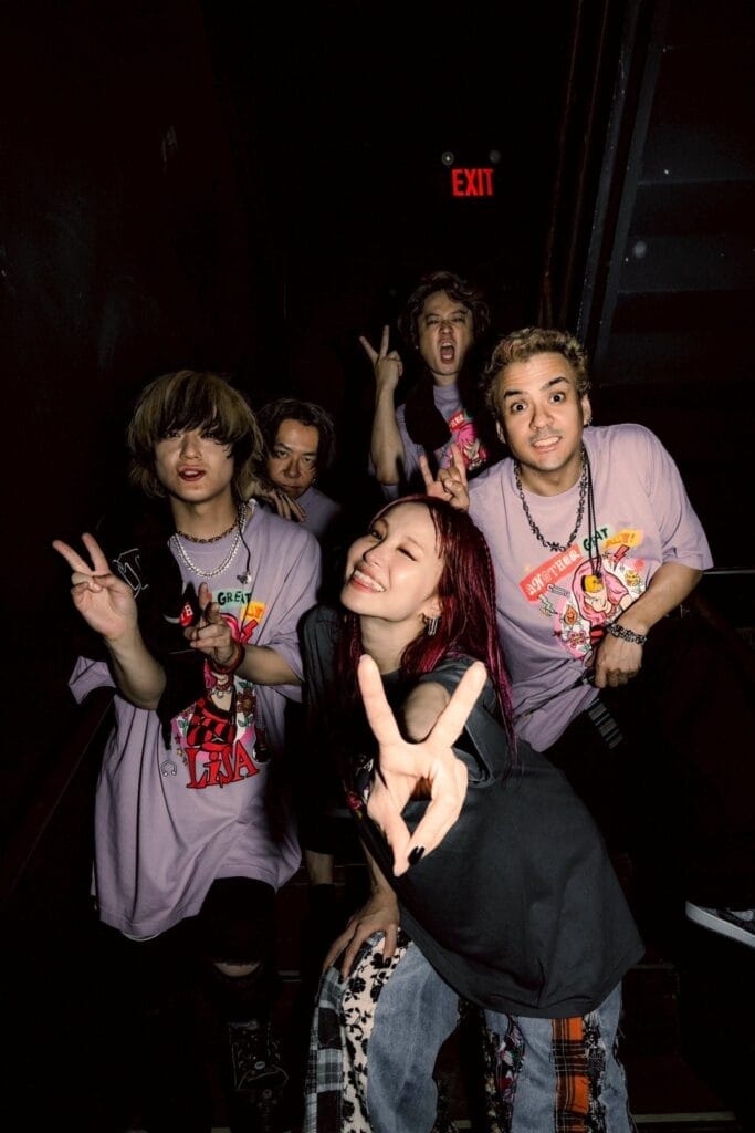 Photograph of LiSA smiling as she flashes a peace sign at the camera. She's surrounded by members of the band, who are dressed in tour T-shirts.
