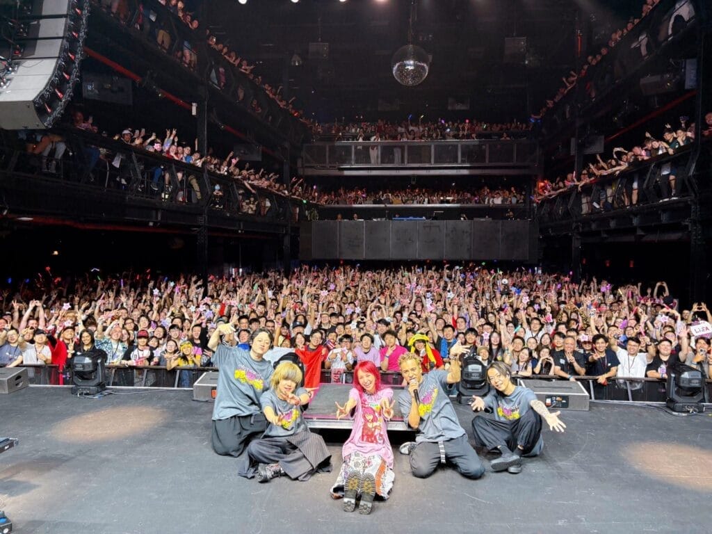 Photograph of LiSA sitting onstage, cross-legged, smiling as she's dressed in a T-shirt and colorful pants. The entire audience is visible behind her.