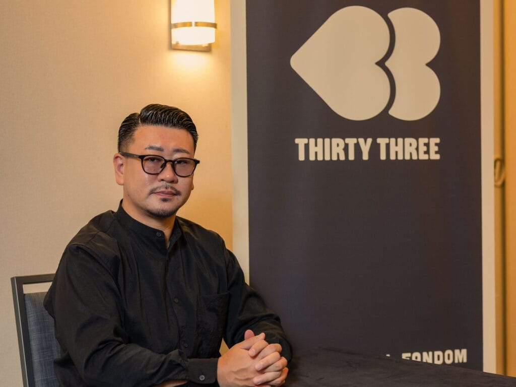 Photograph of 33 Inc. President Kuji Kubota siting in front of a sign with the company's logo