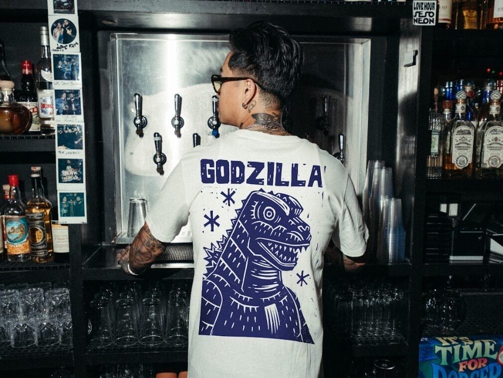 Photograph of artist Attack Peter, whose back is turned to the camera. His shirt features a stylized illustration of Godzilla on the back.